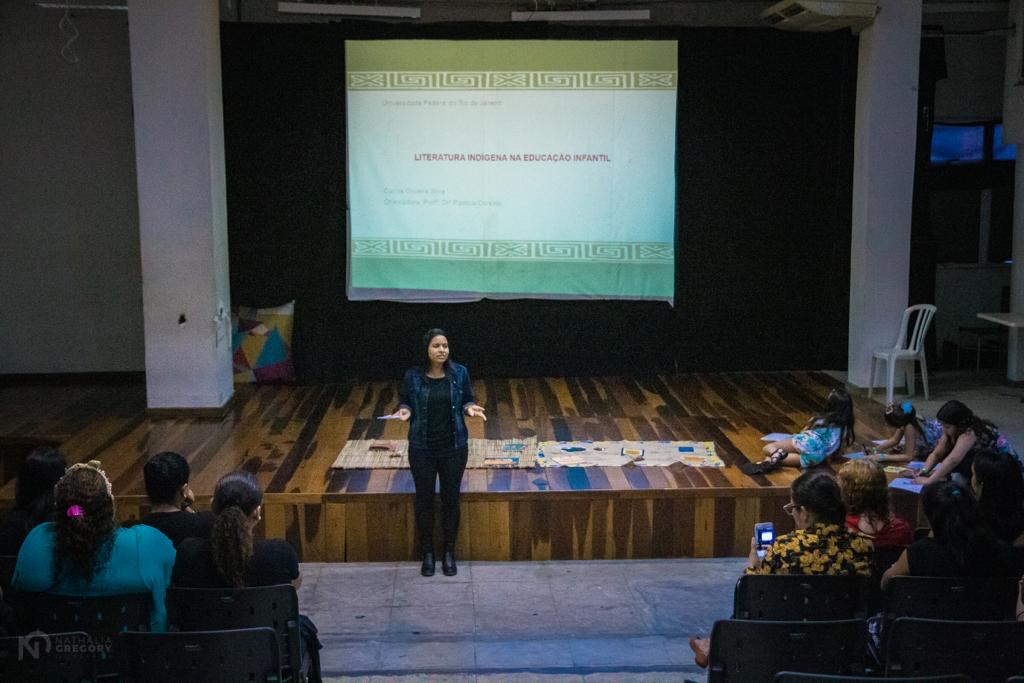 Sala de apresentações de uma biblioteca, com um palco em que uma mulher está de pé proferindo uma palestra, com uma tela branca projetando slides.